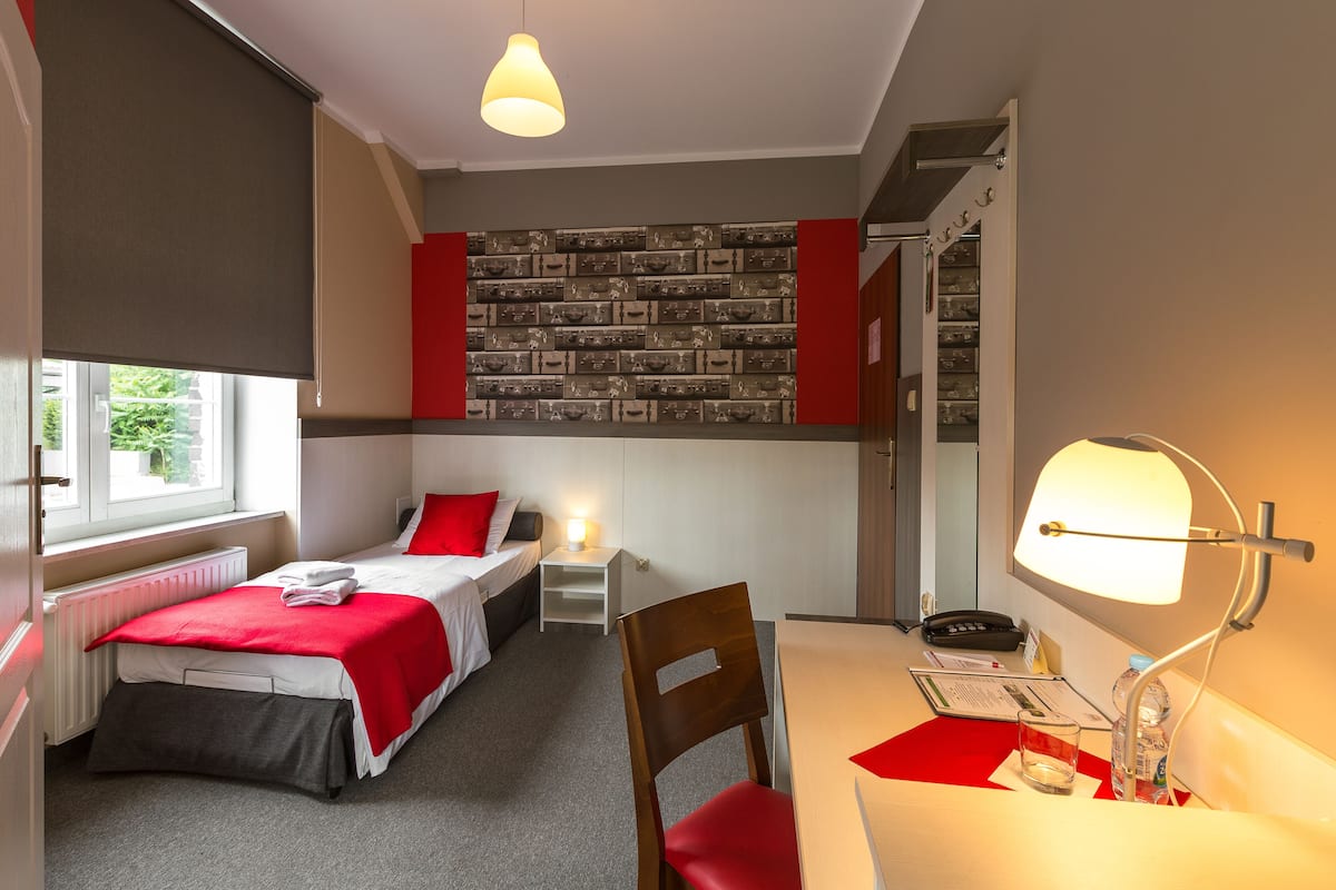 A compact hotel room with a single bed, a desk with a lamp, a chair, a small table, and a red and white color scheme.