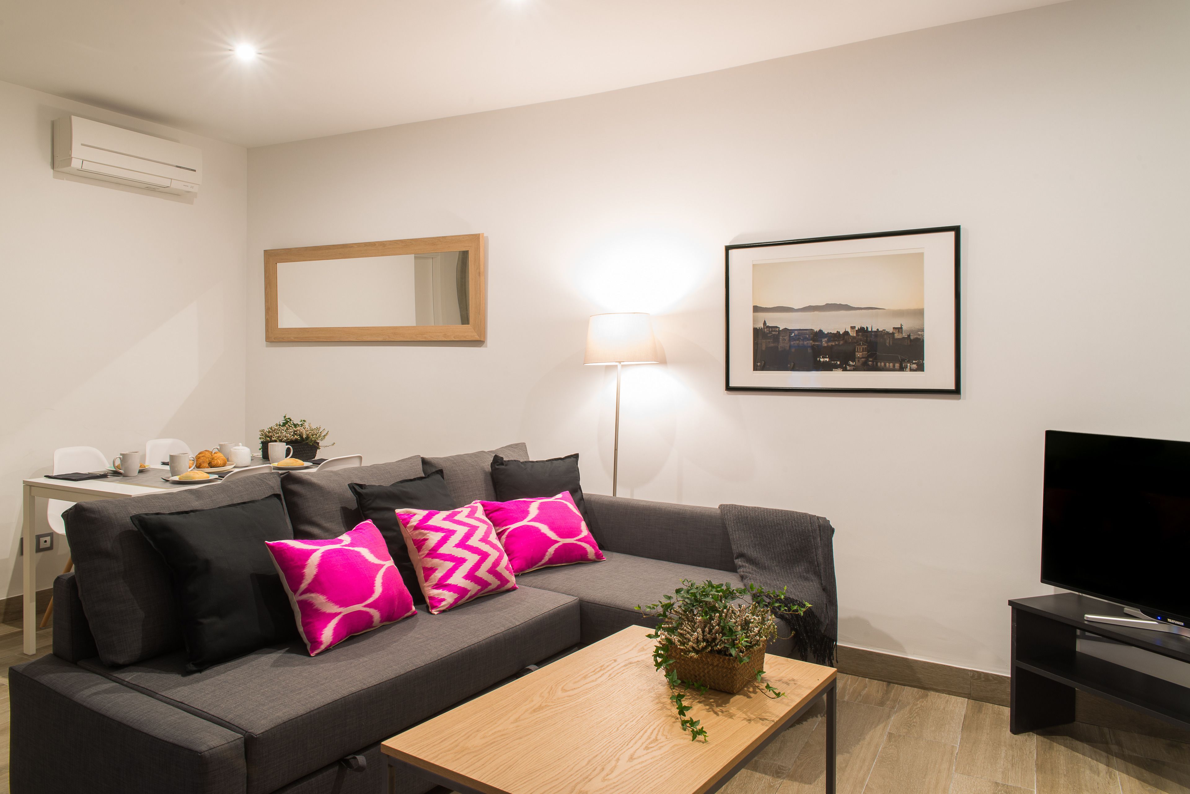 Un salon moderne avec un canapé gris, une table basse en bois, un téléviseur à écran plat, un tableau encadré représentant un paysage urbain, une lampe sur pied et une petite table sur laquelle se trouvent une plante et une théière.