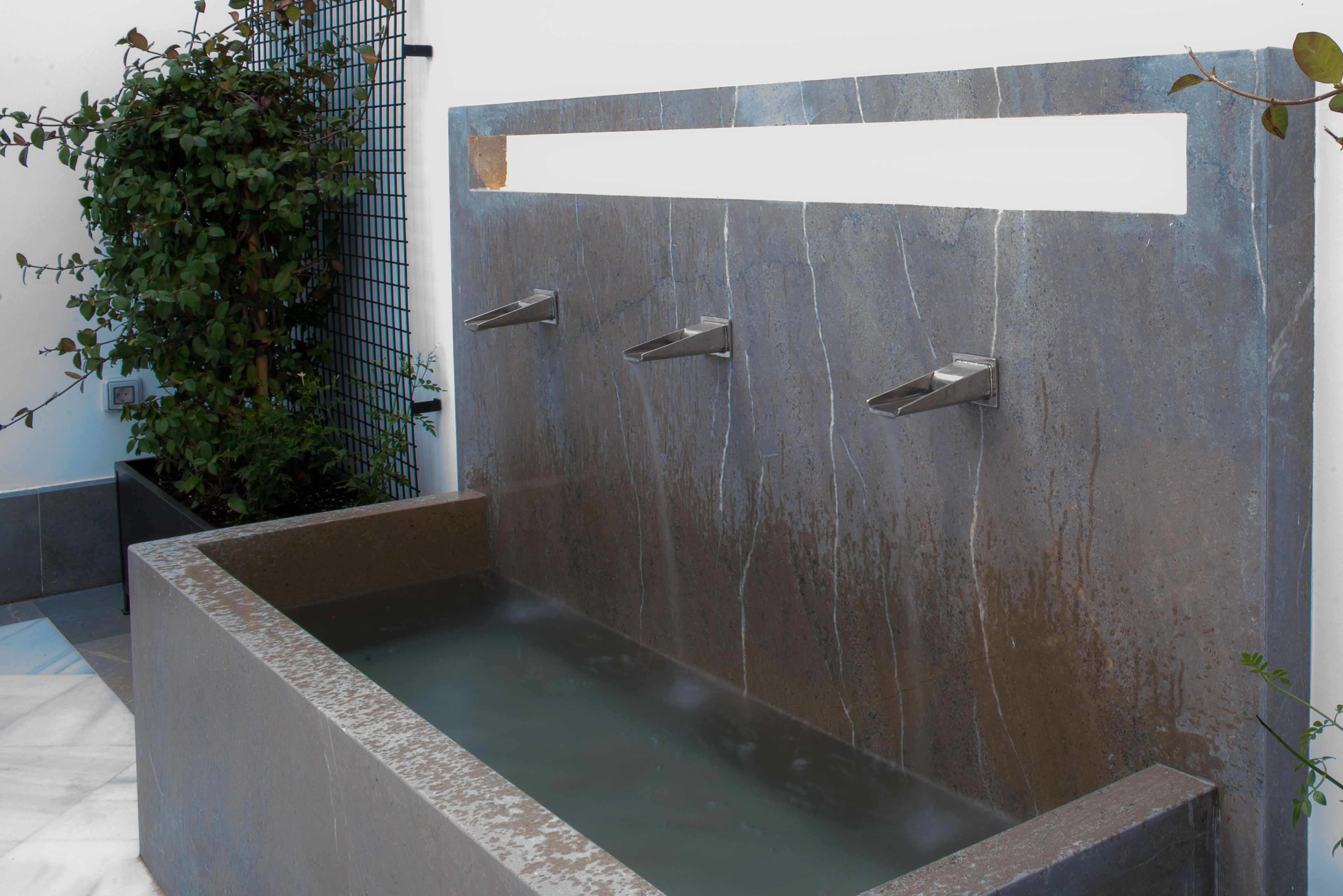 A rectangular stone bathtub with three faucets, a white rectangular panel, and a potted plant beside it.