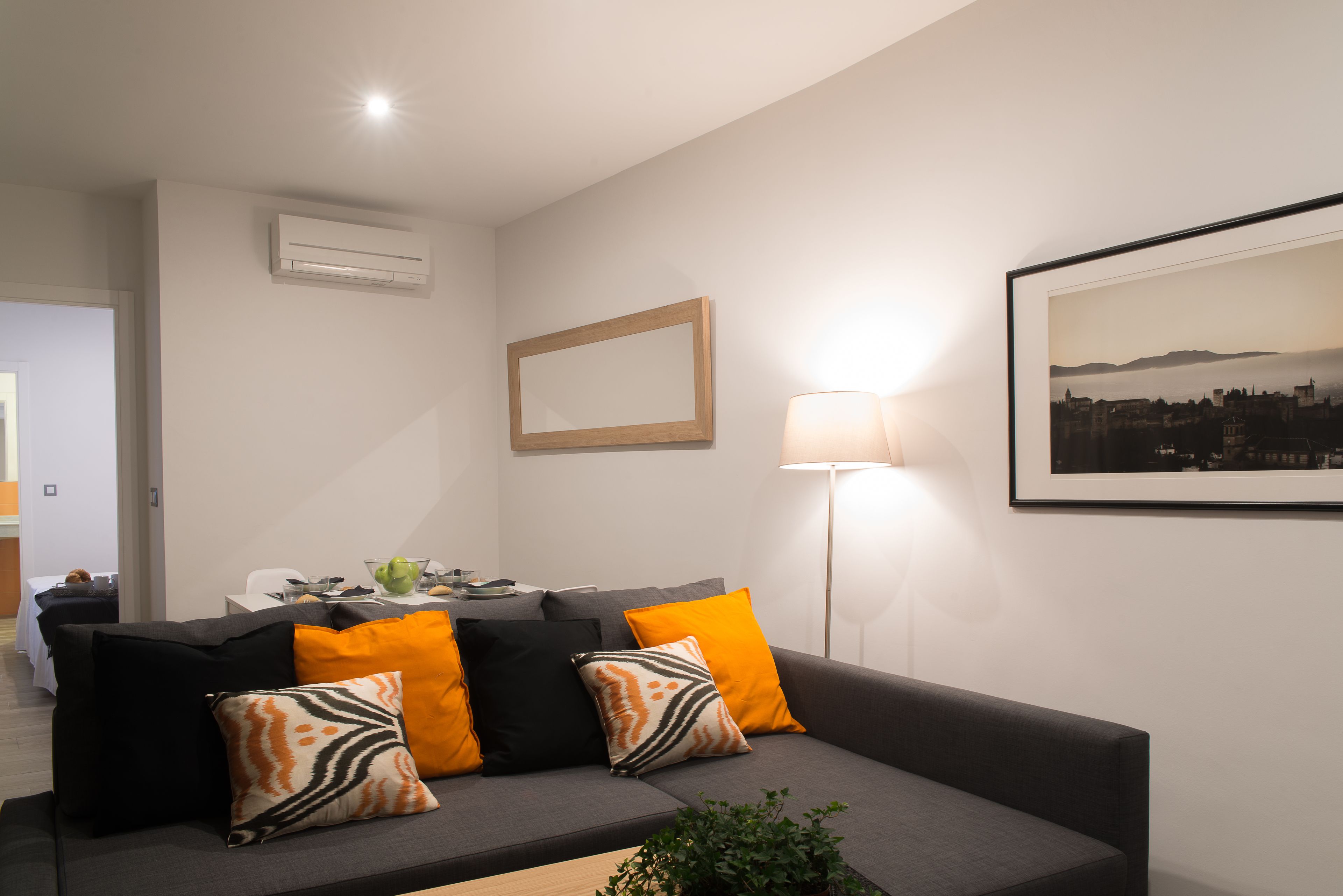 En moderne stue med en grå sofa, orange og sorte puder, et billede i ramme på væggen og en gulvlampe.