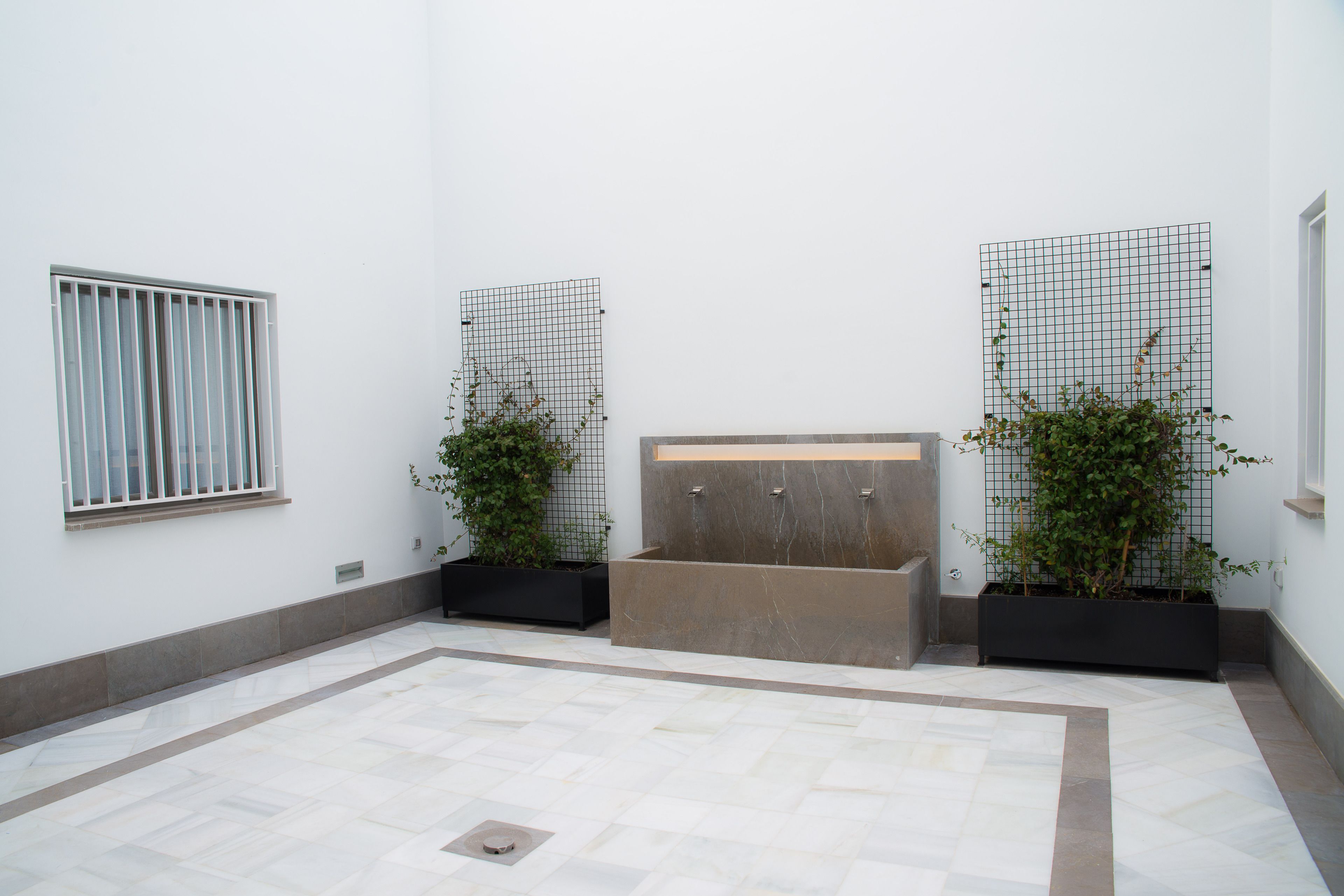 Un espacio interior moderno con suelo de baldosas, una fuente central y dos plantas en macetas.