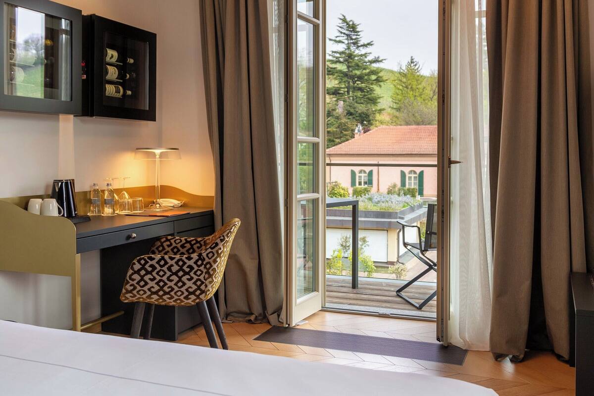 Et hotelværelse med seng, skrivebord med stol, minibar og udsigt ud.