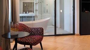 Habitación de hotel con diseño moderno, que incluye un espejo grande, cabina de ducha de vidrio y una zona de estar con sillas estampadas.
