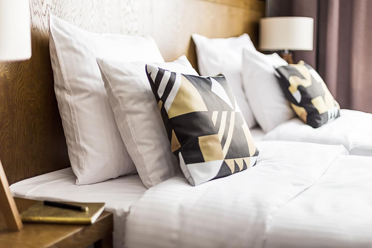 A hotel room with two beds, each with white bedding and decorative pillows. There is a wooden headboard, a bedside table with a lamp, and a book on the table.