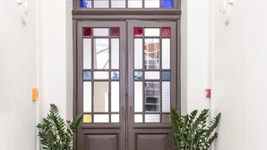 Puertas dobles con paneles de vitral, flanqueadas por plantas en macetas, que dan a un pasillo con suelo estampado.
