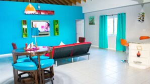 A modern living room with a blue wall, a round dining table with blue chairs, a red sofa, and a TV mounted on the wall. - Casa Tamar Guest Apartments (Kingston)