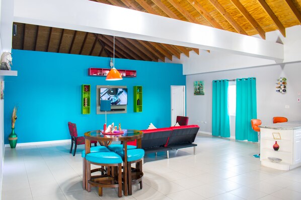A spacious living area with a bright blue wall, wooden ceiling beams, a round dining table with chairs, a sofa, and a television mounted on the wall.