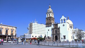 A historic plaza with a prominent church featuring a bell tower and dome, surrounded by modern buildings and a fountain.