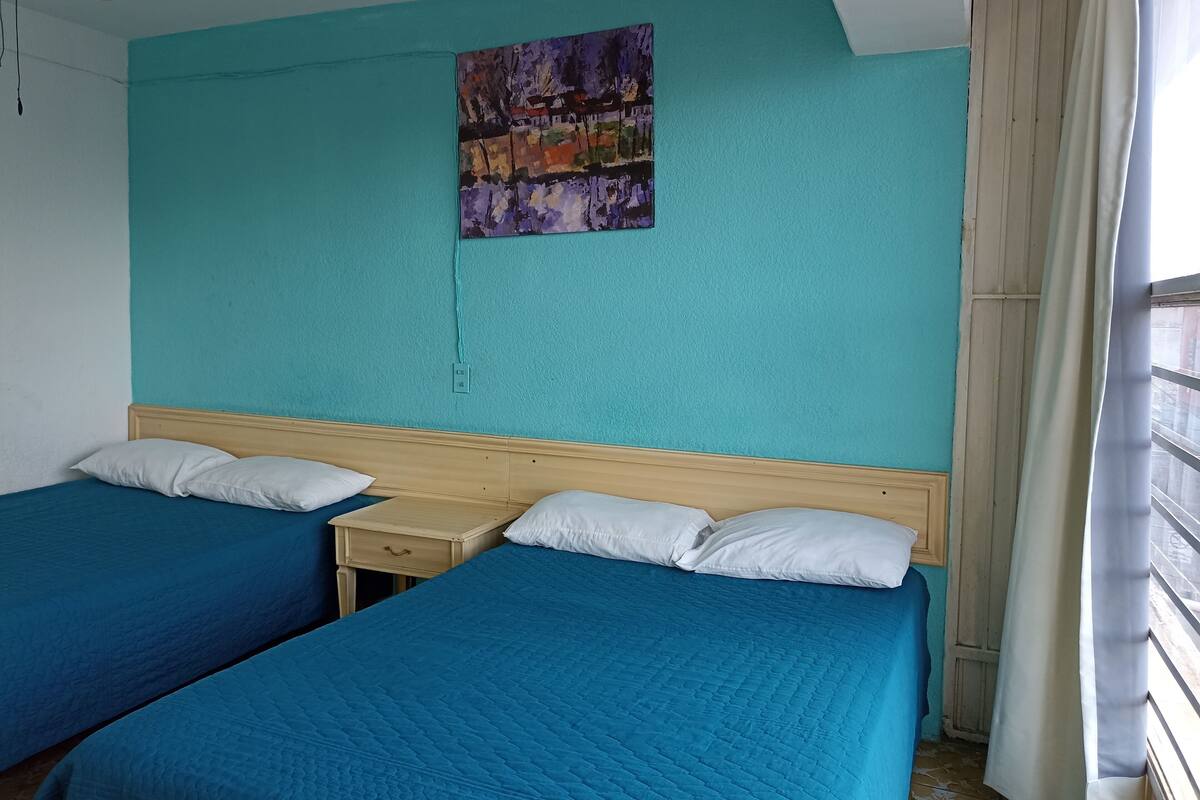 A hotel room with two beds, a small bedside table, a wall painting, and a window with curtains.