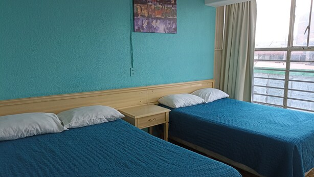 A hotel room with teal walls, two beds with blue bedding, a wooden headboard, a nightstand, a small table, and a window with curtains.
