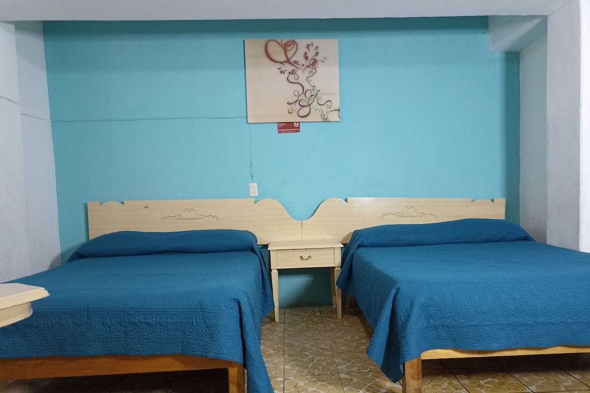 A room with two beds, a small bedside table, a wall-mounted light switch, and a decorative piece above the beds.