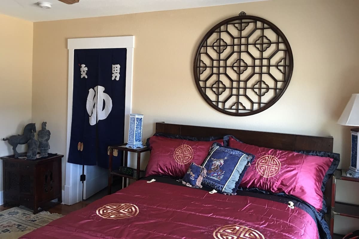 Un dormitorio con cama, almohadones decorativos, un adorno en la pared, cabecera de madera, mesita de noche, lámpara y cómoda.