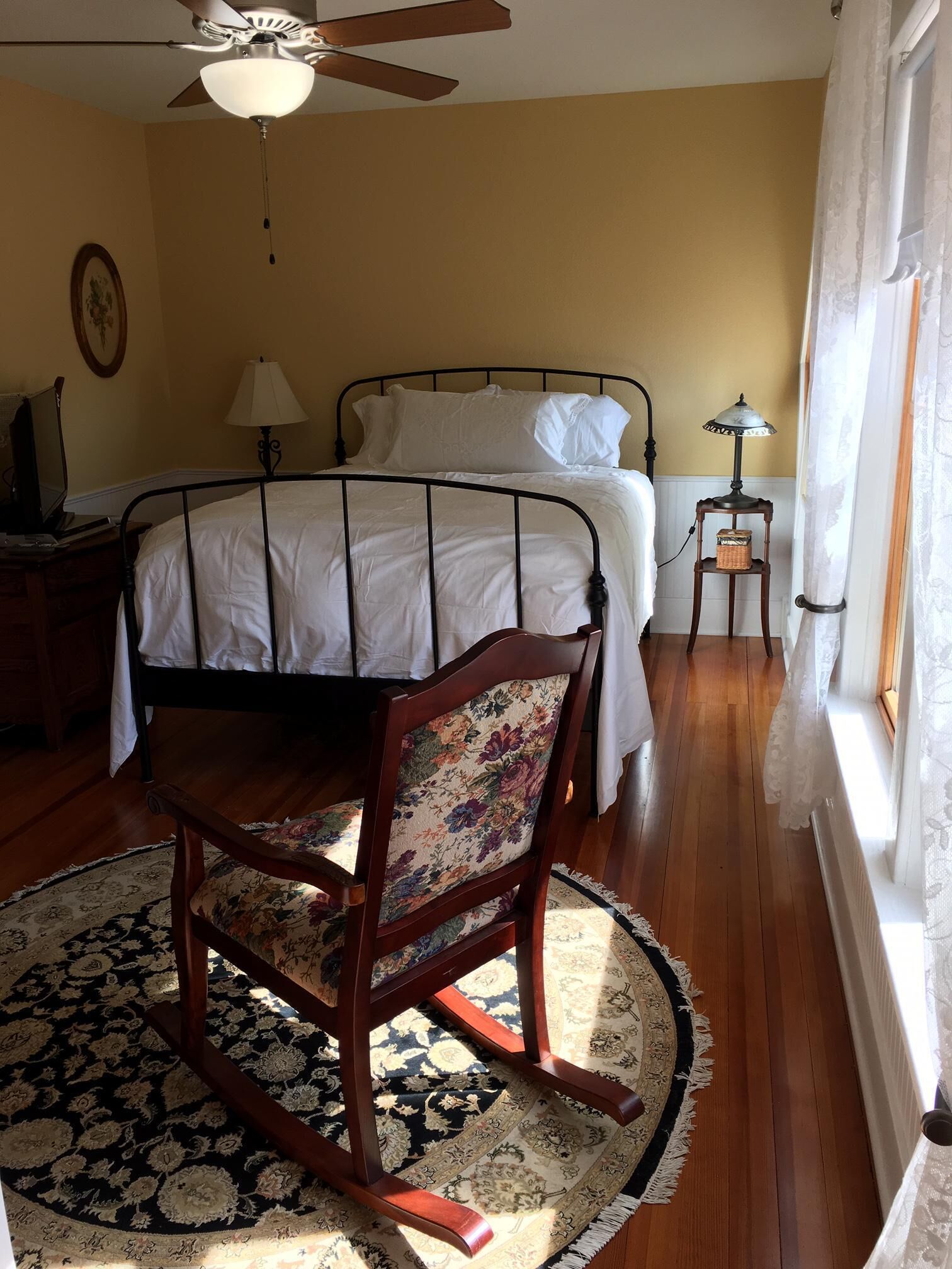 A bedroom with a bed, a wooden chair with floral upholstery, a ceiling fan, a bedside table with a lamp, and a window with curtains.