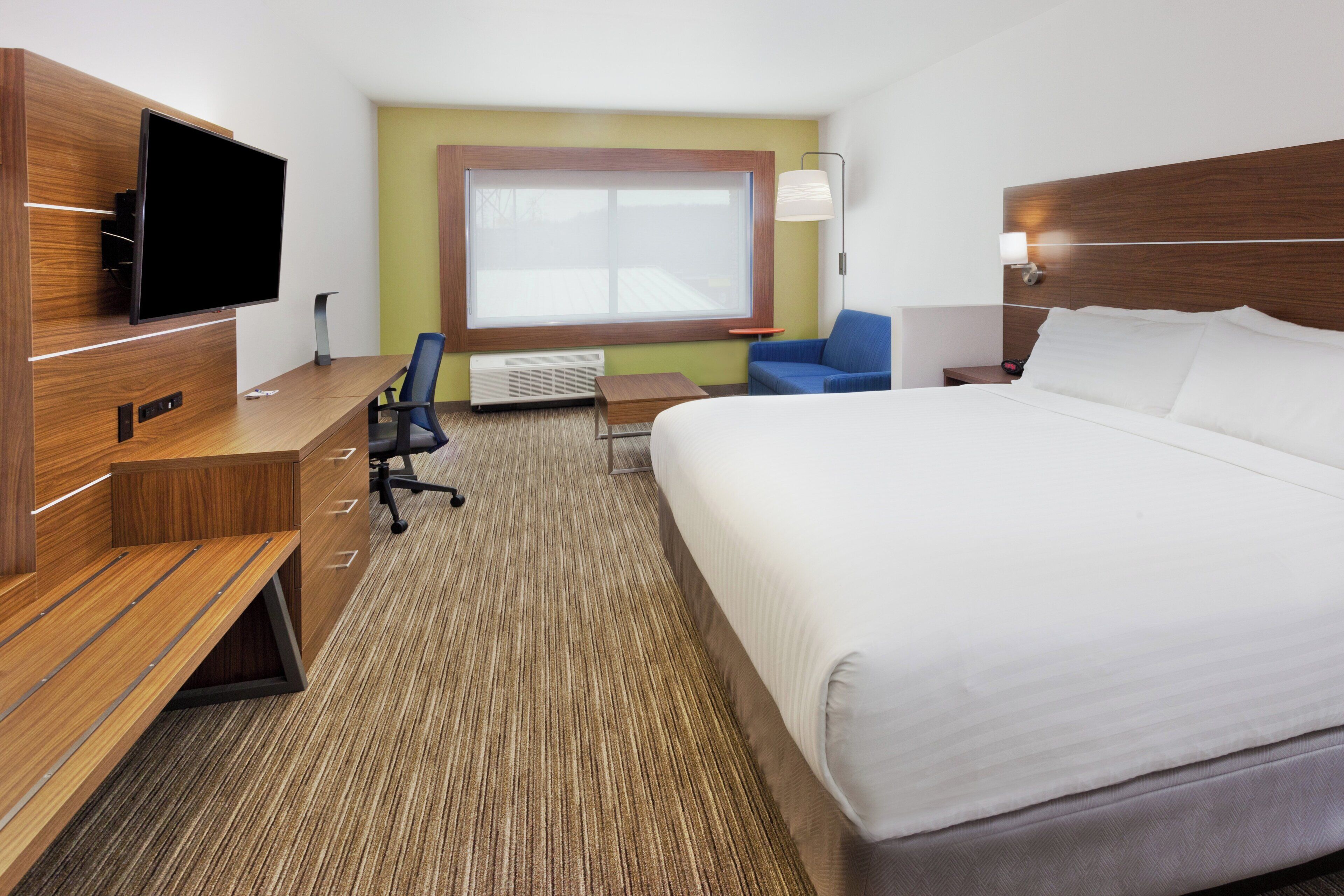 A hotel room with a bed, a desk with a flat-screen TV, a chair, a small table, and a window.