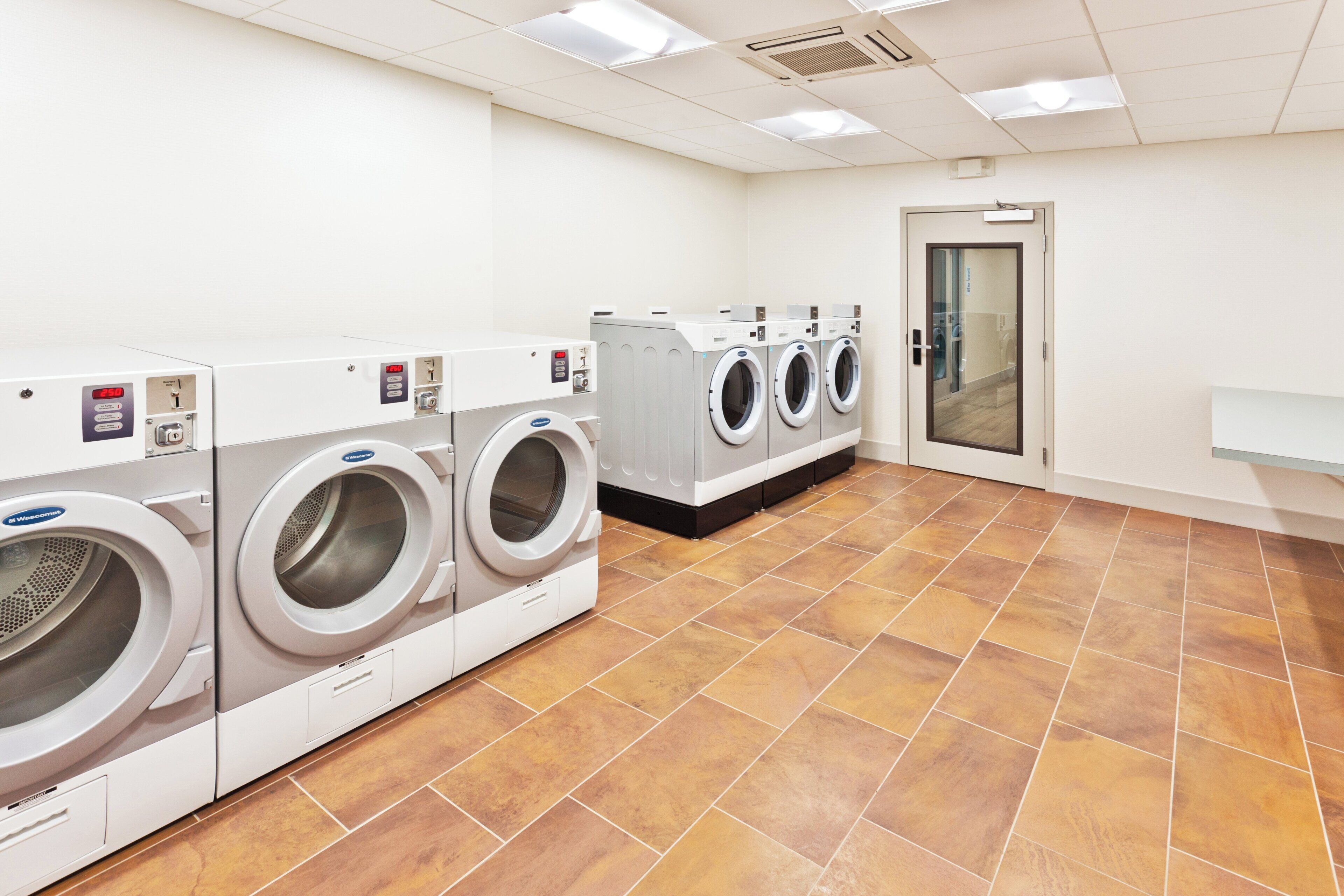 Une buanderie équipée de plusieurs machines à laver et de plusieurs sèche-linge, avec un sol carrelé et une porte donnant sur une autre pièce.