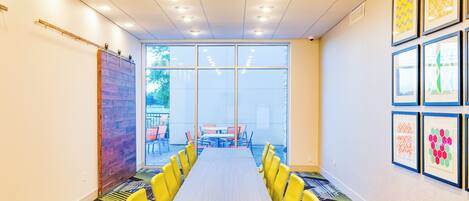 A conference room with a long table, yellow chairs, and a view of an outdoor area with tables and chairs.