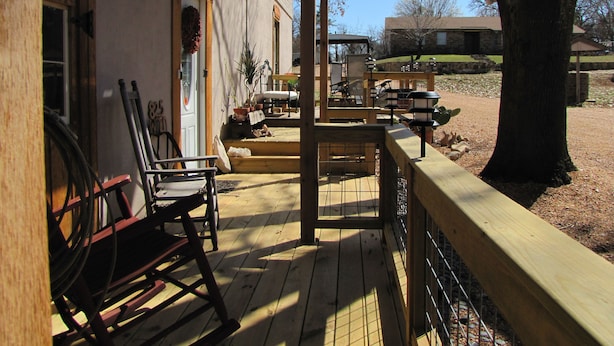Una terraza de madera con mobiliario exterior, una zona cubierta con sillas y una vista de un edificio al fondo.