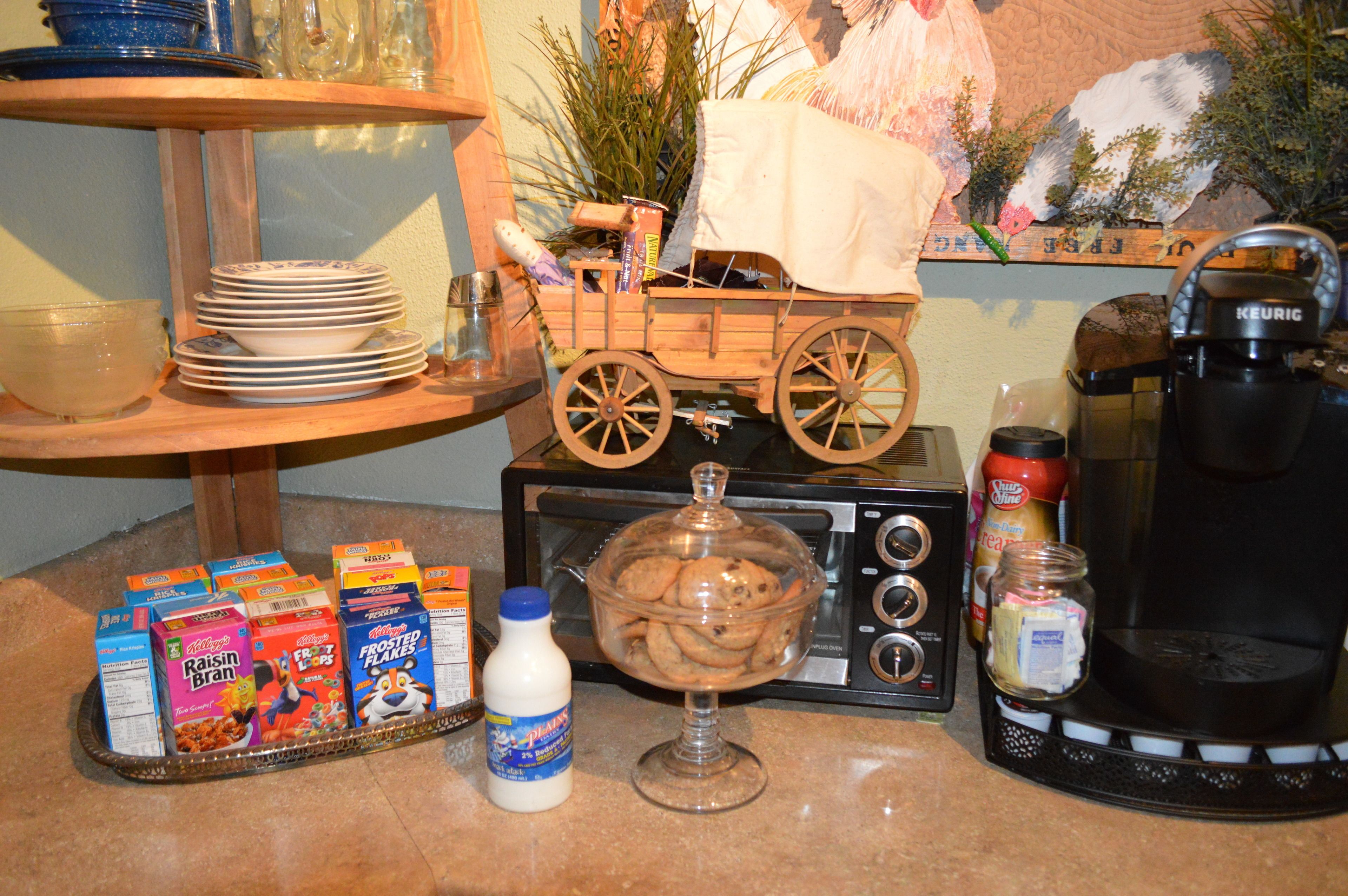 Una encimera de cocina con cajas de cereal, una botella de leche, un tarro de vidrio, una cafetera y un carrito de madera con una manta.