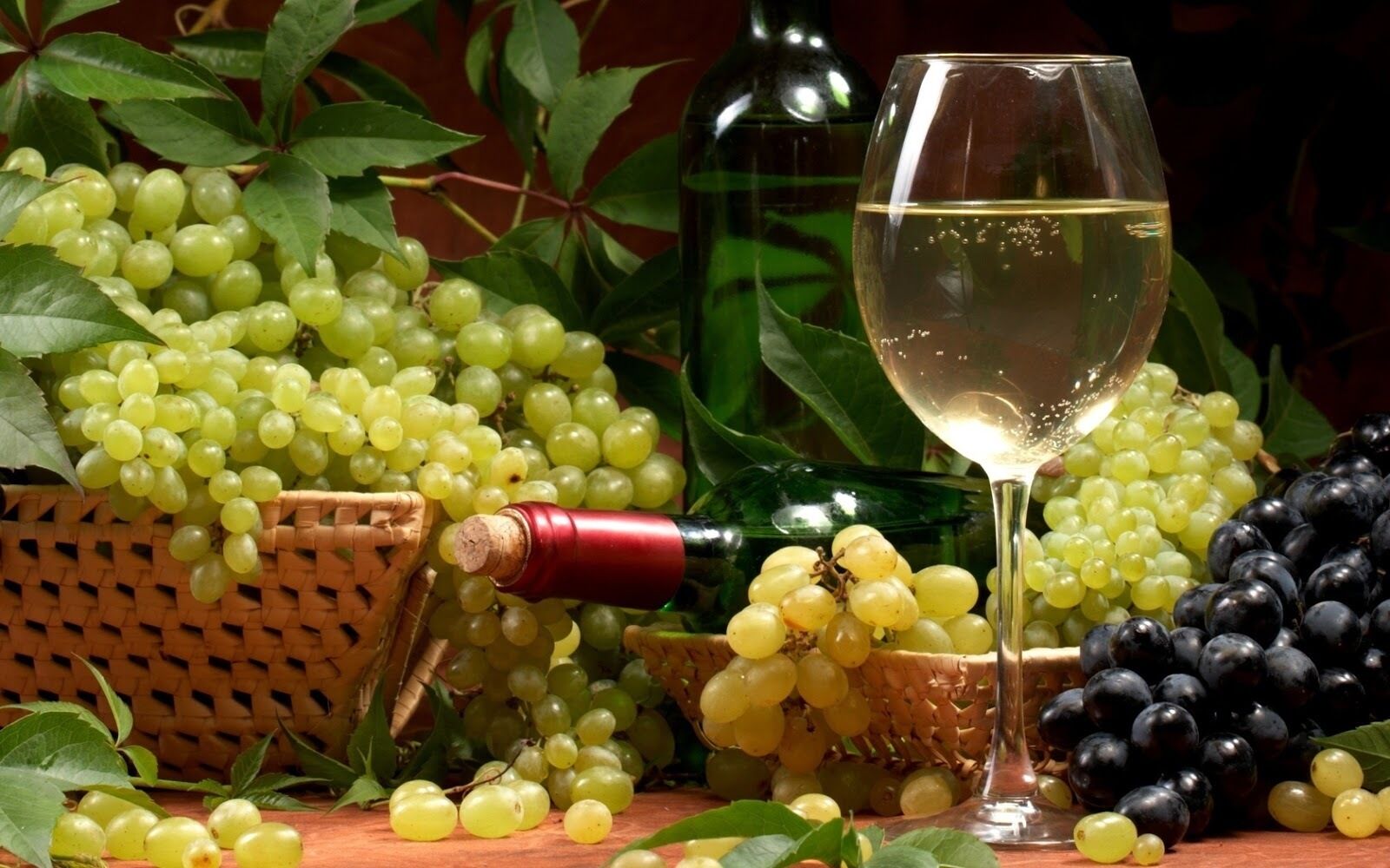 A table with a bottle of white wine, a glass of wine, and baskets of green and black grapes.