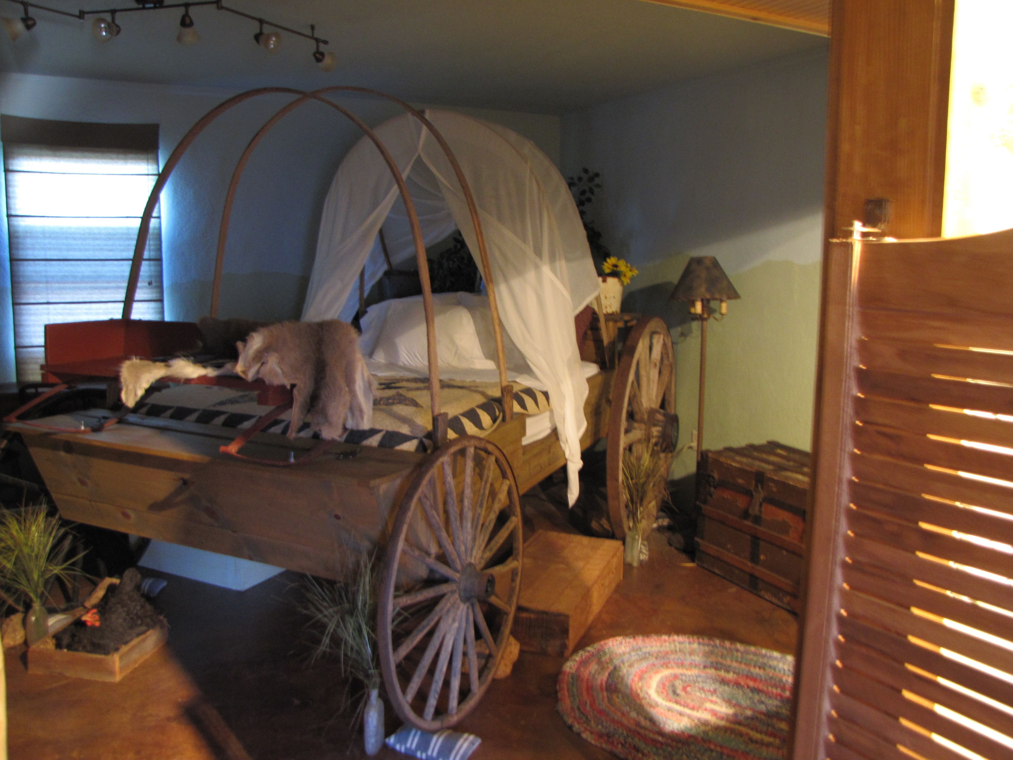 A rustic room with a bed featuring a canopy, a wooden cart-like structure, and a vintage lamp.