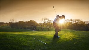 Una persona en un campo de golf, con un palo de golf, sol y árboles al fondo.
