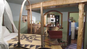 Una habitación rústica con una cama a dosel, una silla de madera, un carro de madera y una zona de cocina al fondo.