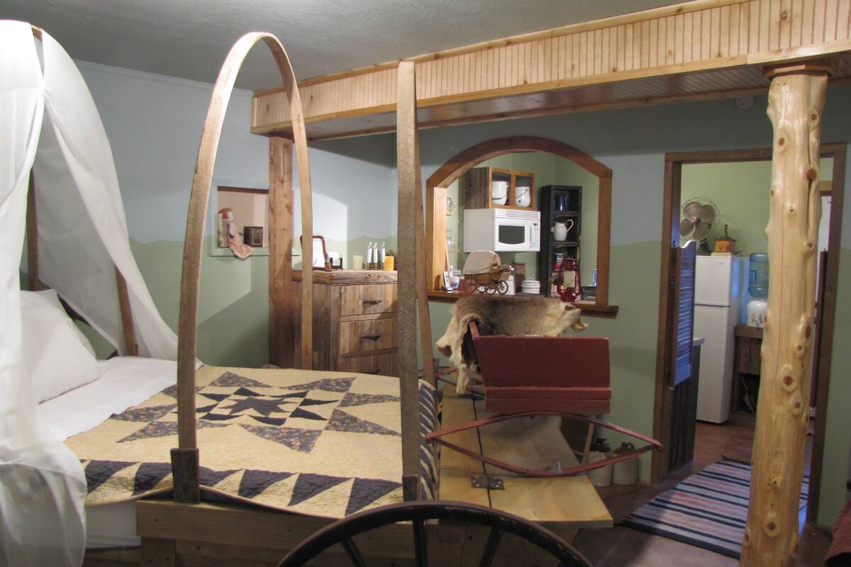 A rustic room with a canopy bed, a wooden chair, a wooden cart, and a kitchen area in the background.