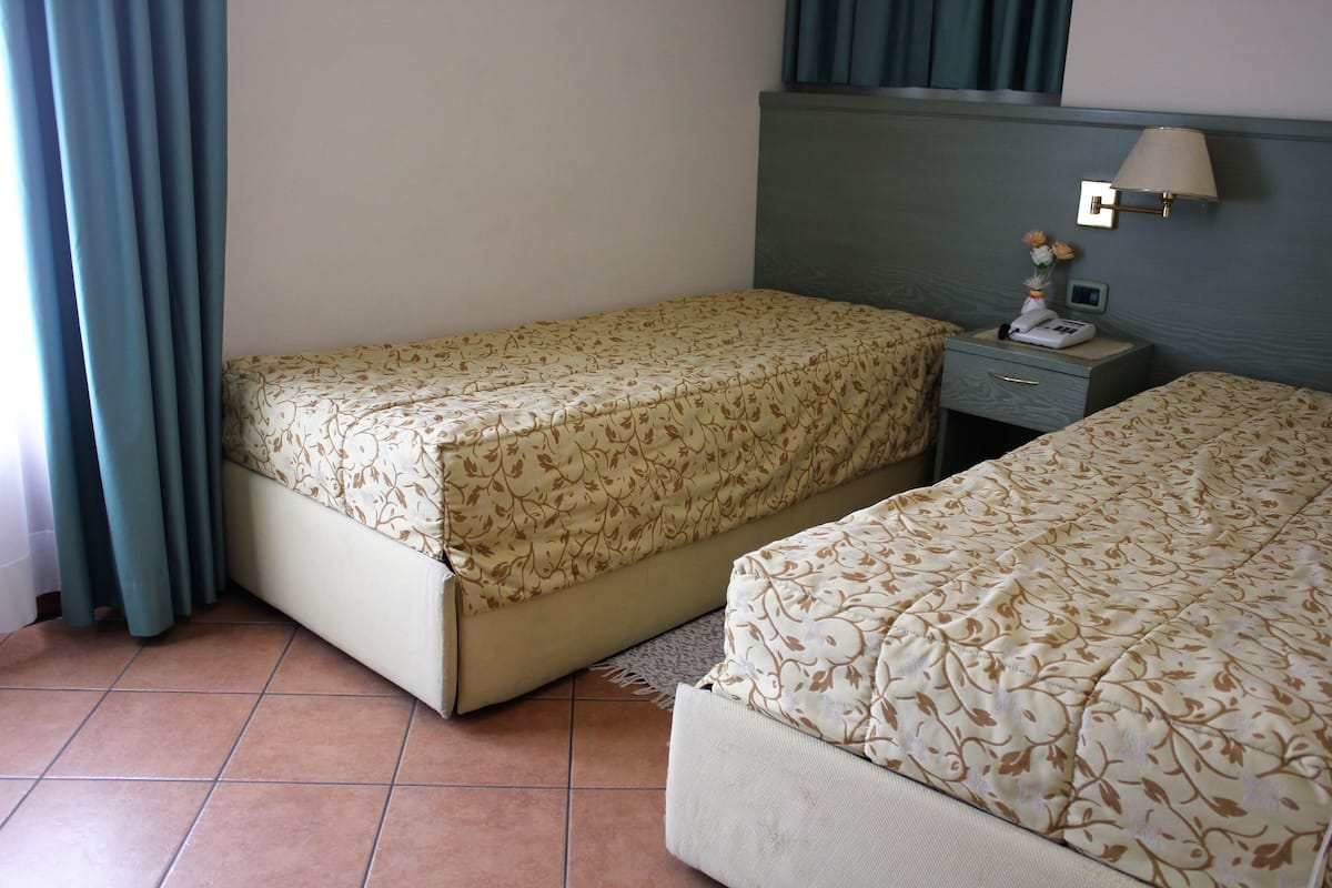A hotel room with two beds, a nightstand with a lamp, and a window with curtains.