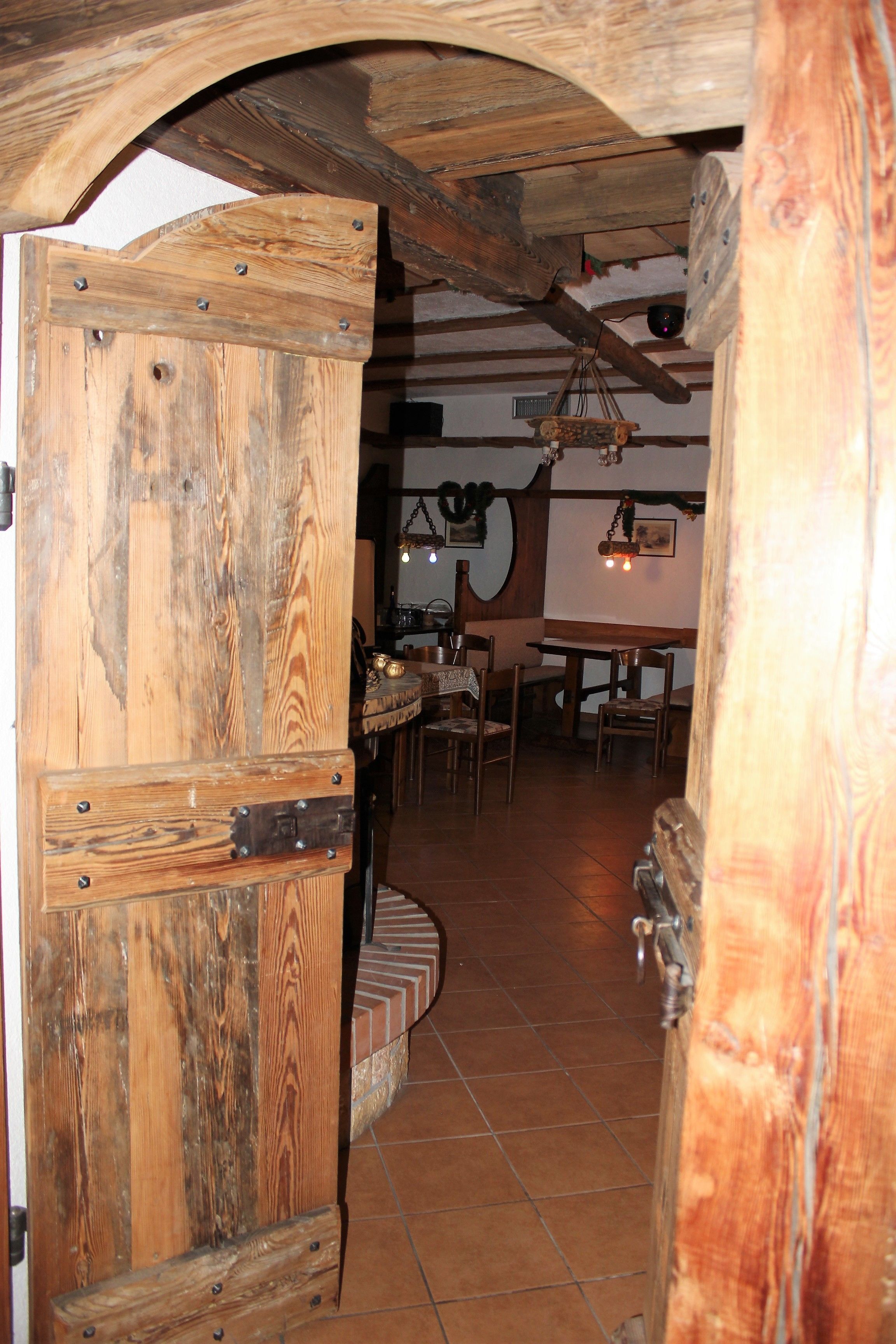 Une porte en bois rustique s’ouvre sur un intérieur également rustique, avec des poutres apparentes, un sol carrelé et un coin repas comprenant des chaises et une table.