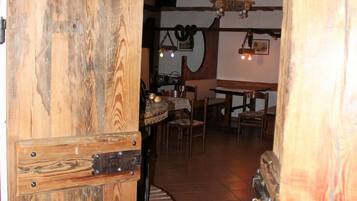 Une porte en bois rustique s’ouvre sur un intérieur également rustique, avec des poutres apparentes, un sol carrelé et un coin repas comprenant des chaises et une table.