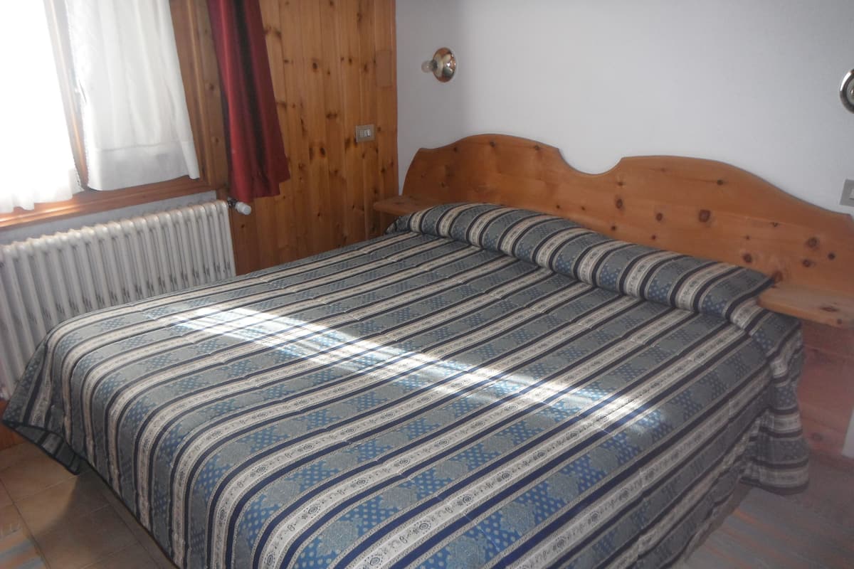 A bedroom with a wooden headboard, a striped bedspread, a radiator, and a window with curtains.