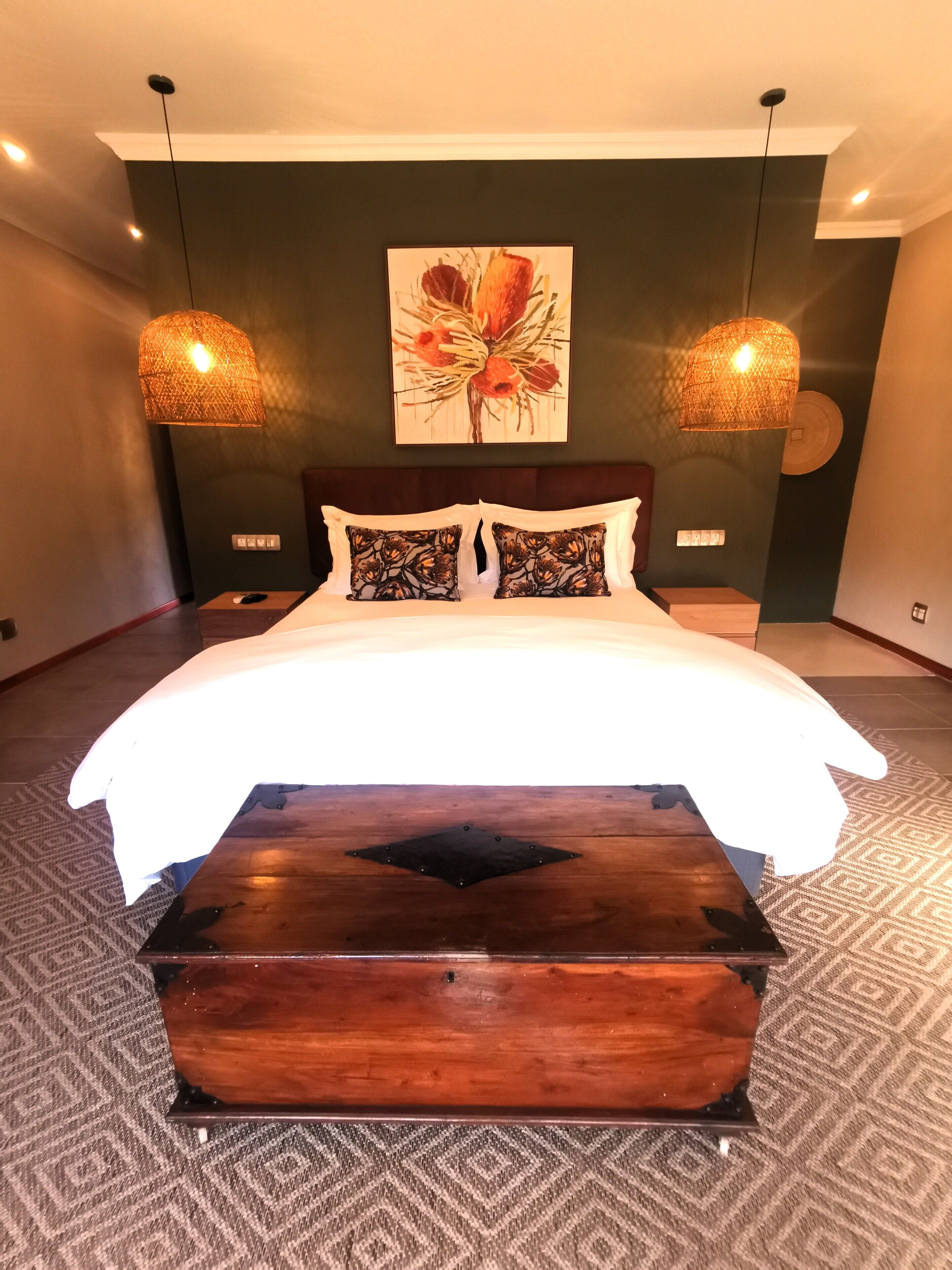 Um quarto moderno com uma cama grande, duas luminárias fixadas na parede, um baú de madeira e uma pintura floral na parede.