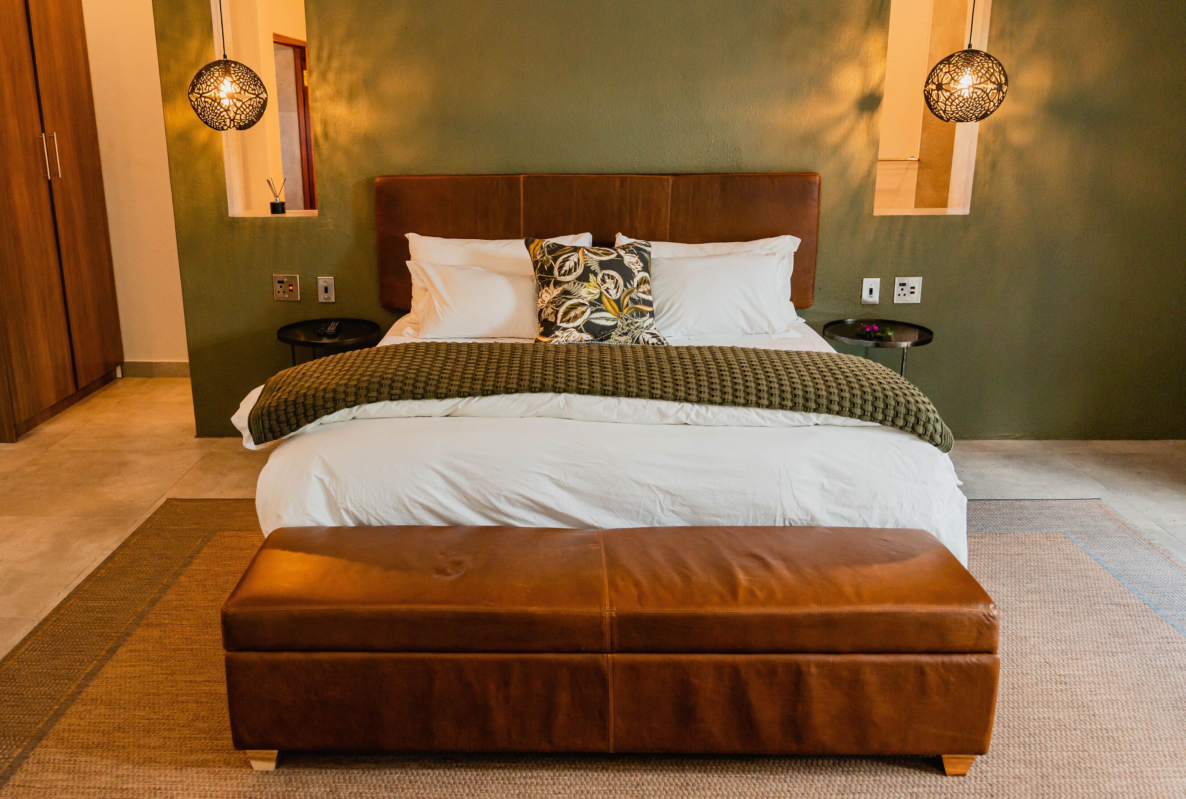 A neatly made bed with a brown leather ottoman in front, a wooden headboard, and two bedside tables with lamps.