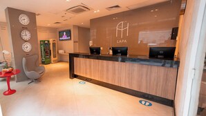 A hotel reception area with a wooden reception desk, a television mounted on the wall, and clocks displaying different city times. - Rede Andrade Lapa (Rio de Janeiro)