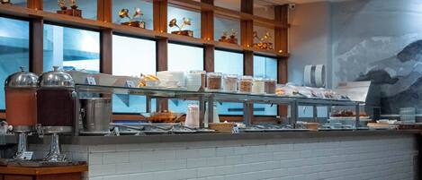 A buffet area with various food items and beverages, wooden shelves with decorative items, and a brick wall.