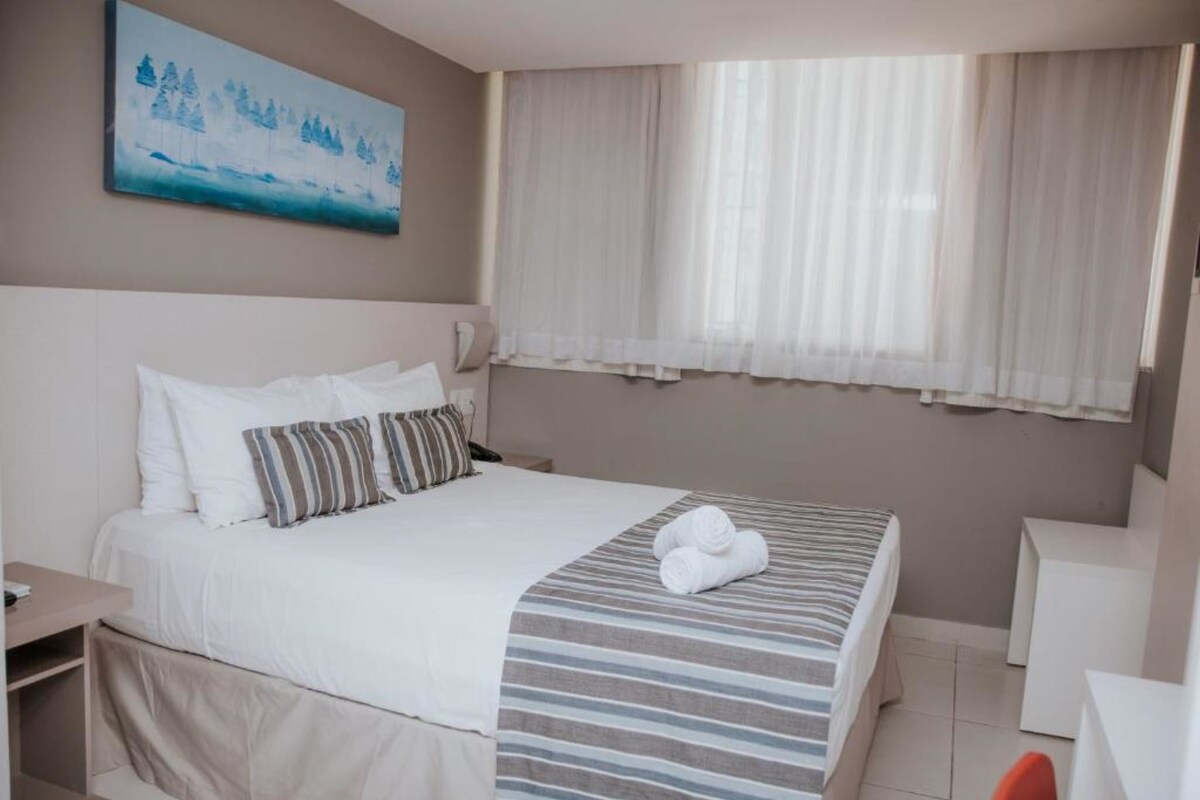 A hotel room with a bed, a bedside table, a chair, and a window with curtains.