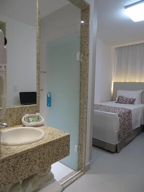 A hotel room with a bathroom featuring a granite countertop sink, a mirror, and a towel rack. There is a bed with a white and patterned bedspread and a headboard with a light fixture. - Rede Andrade Lapa (Rio de Janeiro)