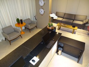 A modern living room with a black kitchen island, a grey sofa, and two armchairs. There are two clocks on the wall, one labeled 'Paris' and the other 'Tokyo'. A small table with a plant and a vase is also visible. - Rede Andrade Lapa (Rio de Janeiro)