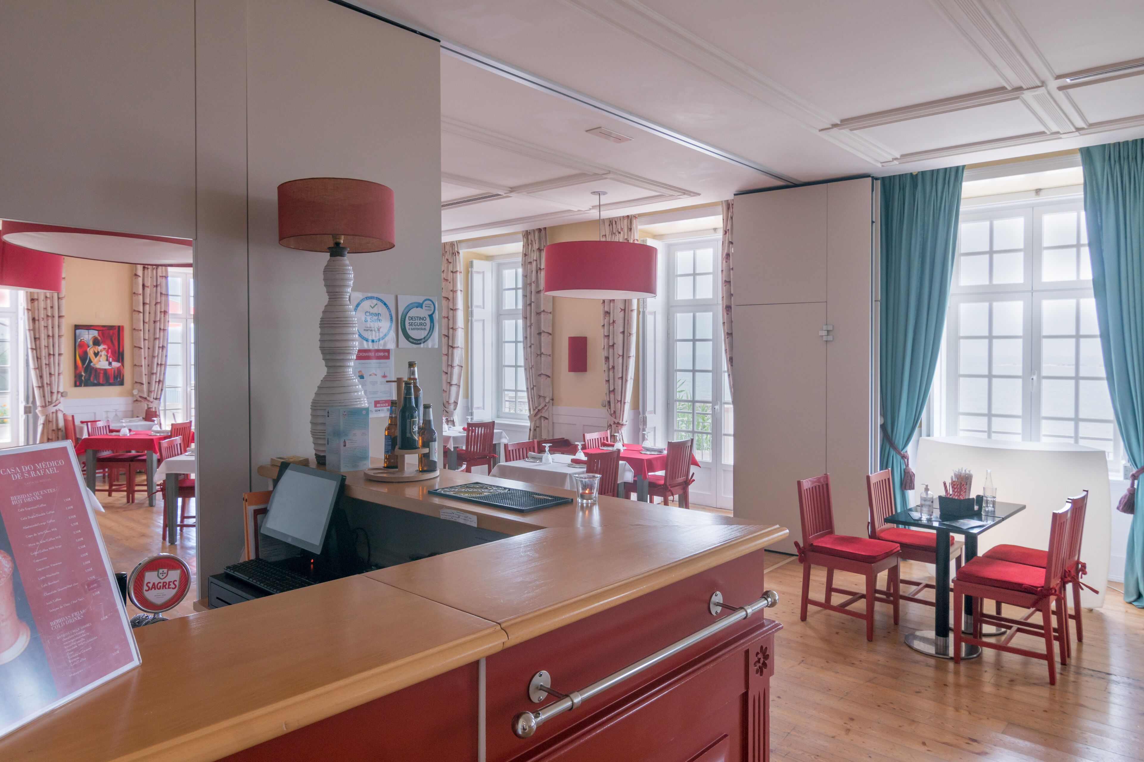 Een restaurantgedeelte in een hotel met houten tafels, rode stoelen en een centraal gelegen balie met een laptop.