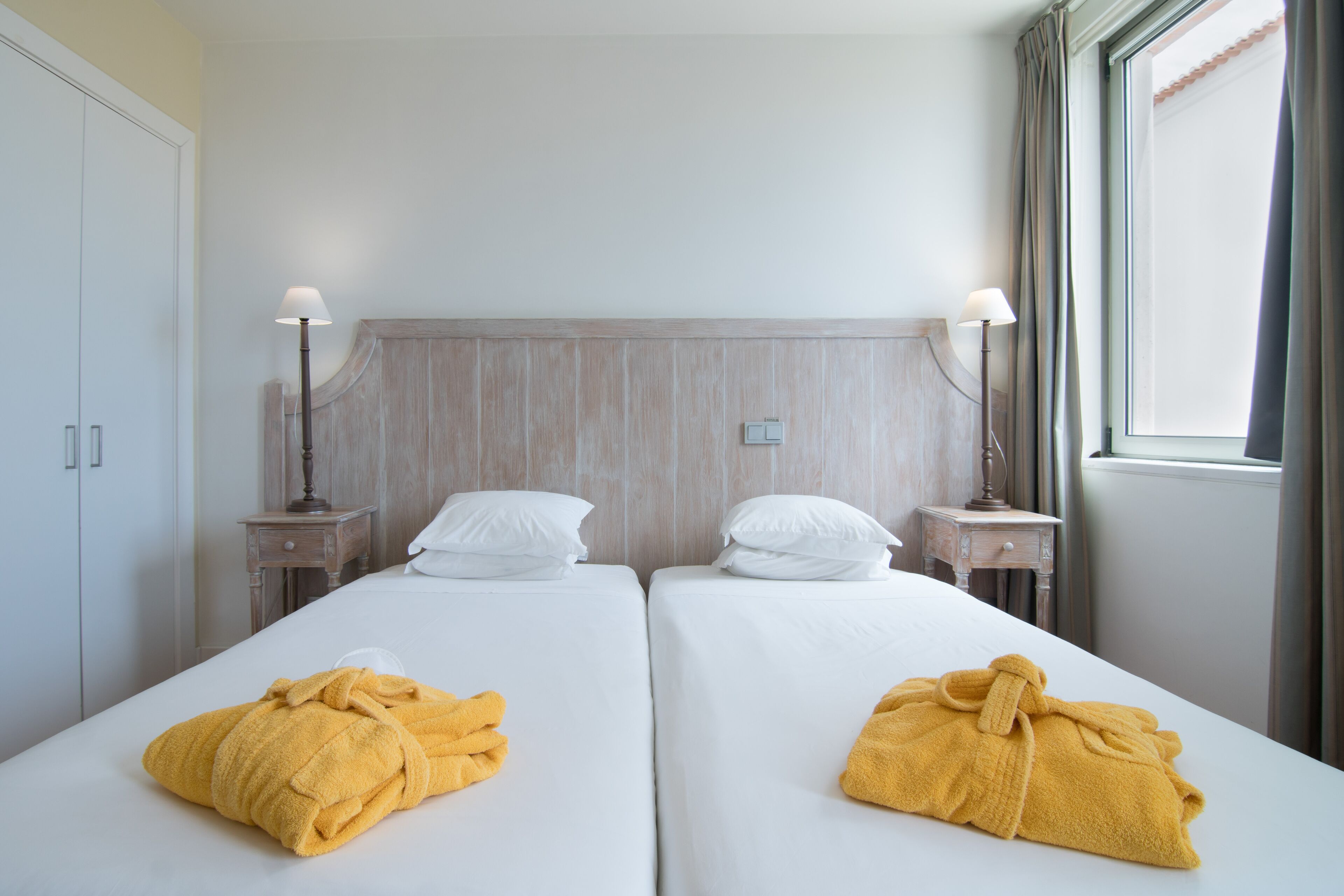 A hotel room with two beds, each with white linens and a yellow towel. There are bedside tables with lamps, a wooden headboard, and a window with curtains.