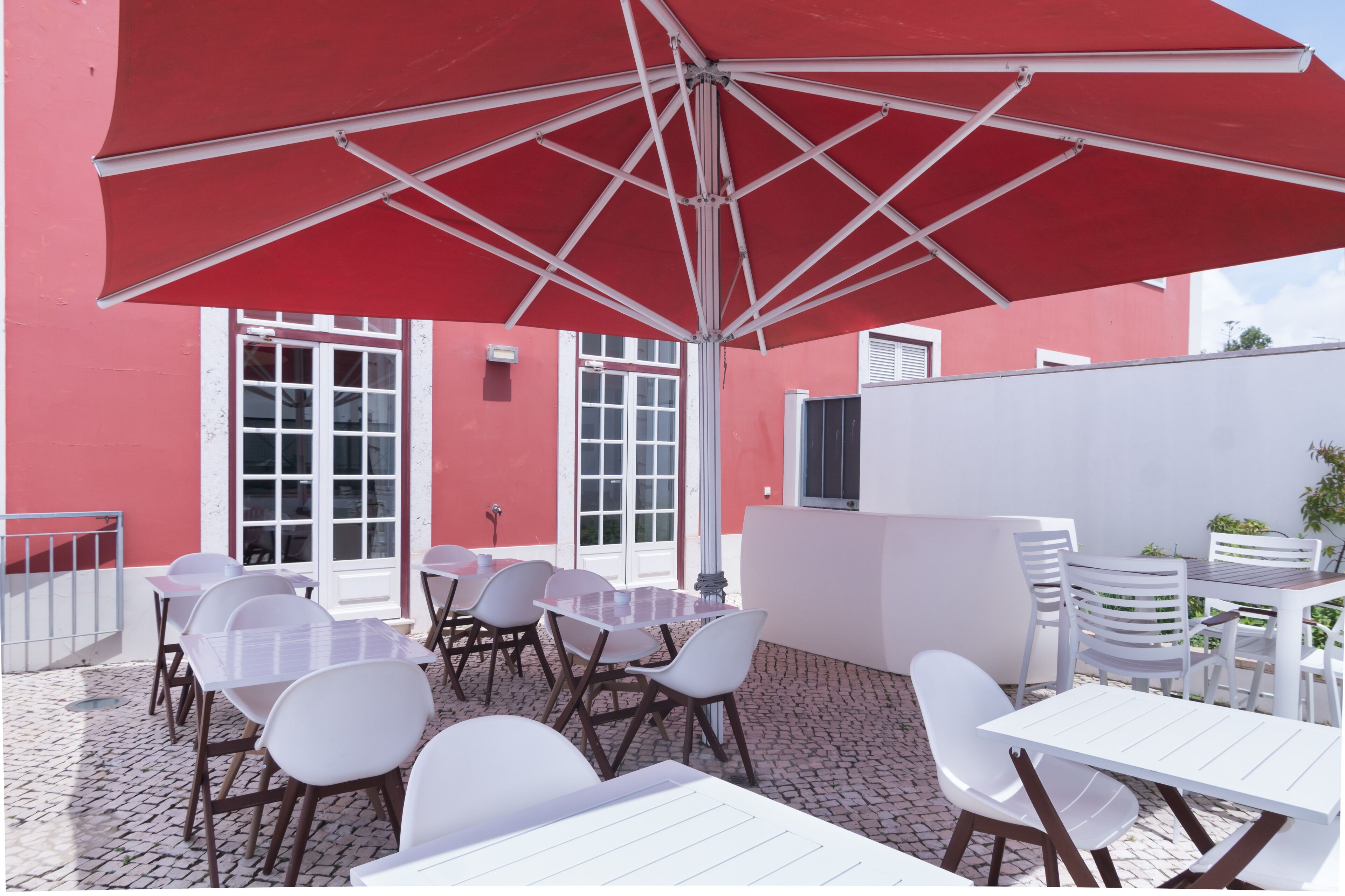 Un patio al aire libre con mesas y sillas blancas, un paraguas rojo y un edificio rojo al fondo.