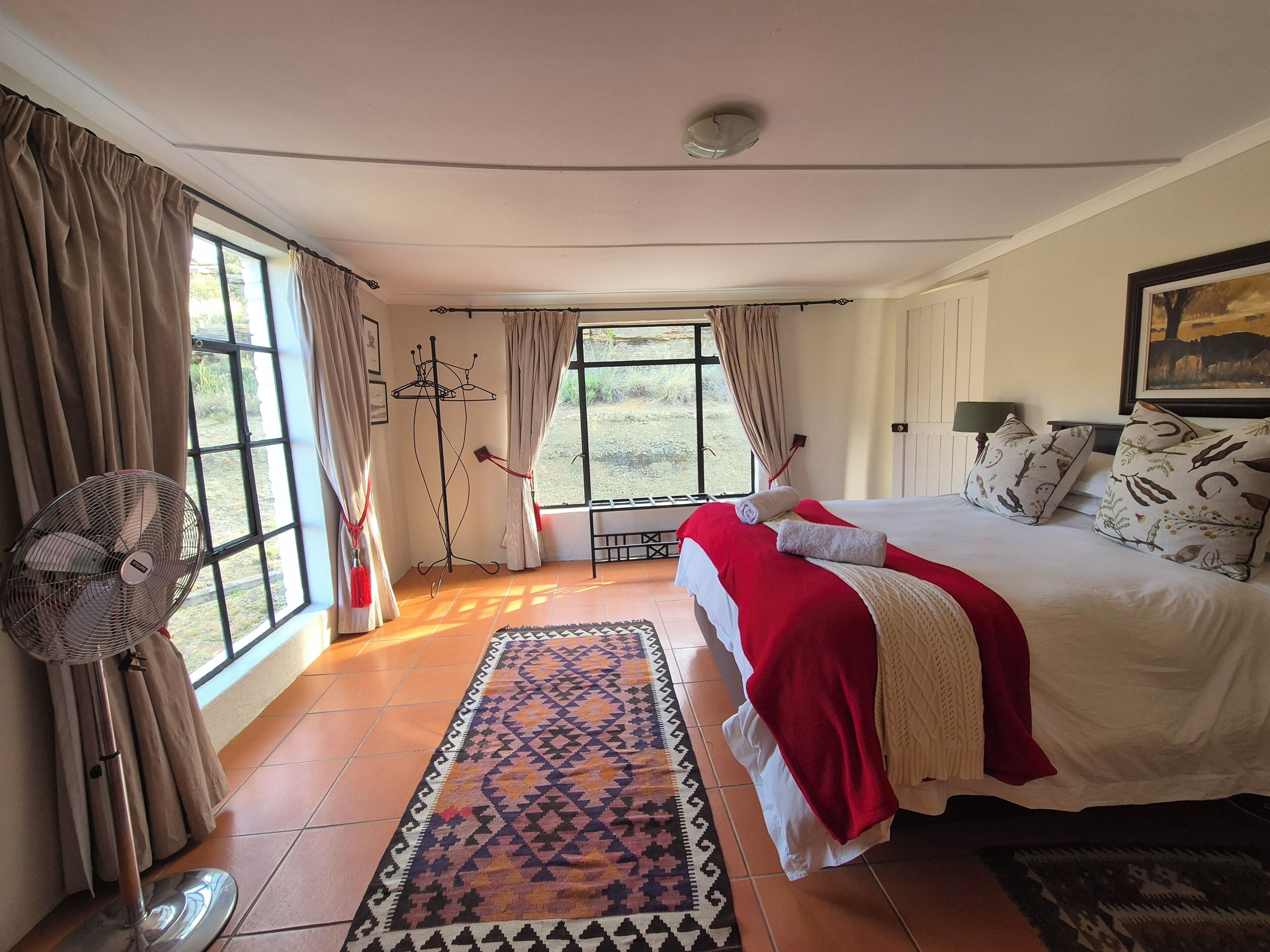 A bedroom with a large bed, a standing fan, a window with curtains, and a patterned rug.