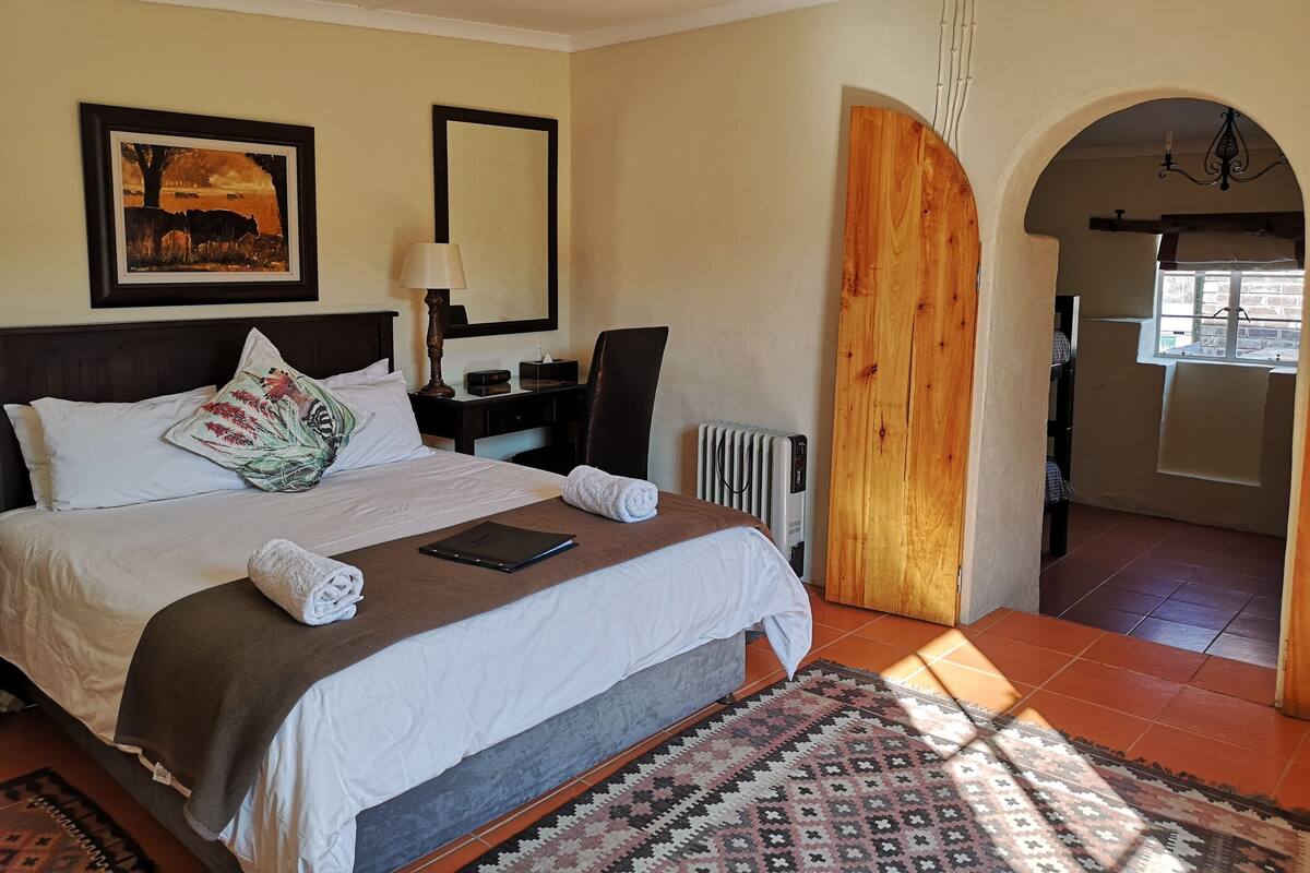 Une chambre d’hôtel avec un grand lit, une table de chevet avec une lampe, un tableau encadré au mur, un radiateur et une porte en arc donnant sur une autre pièce.