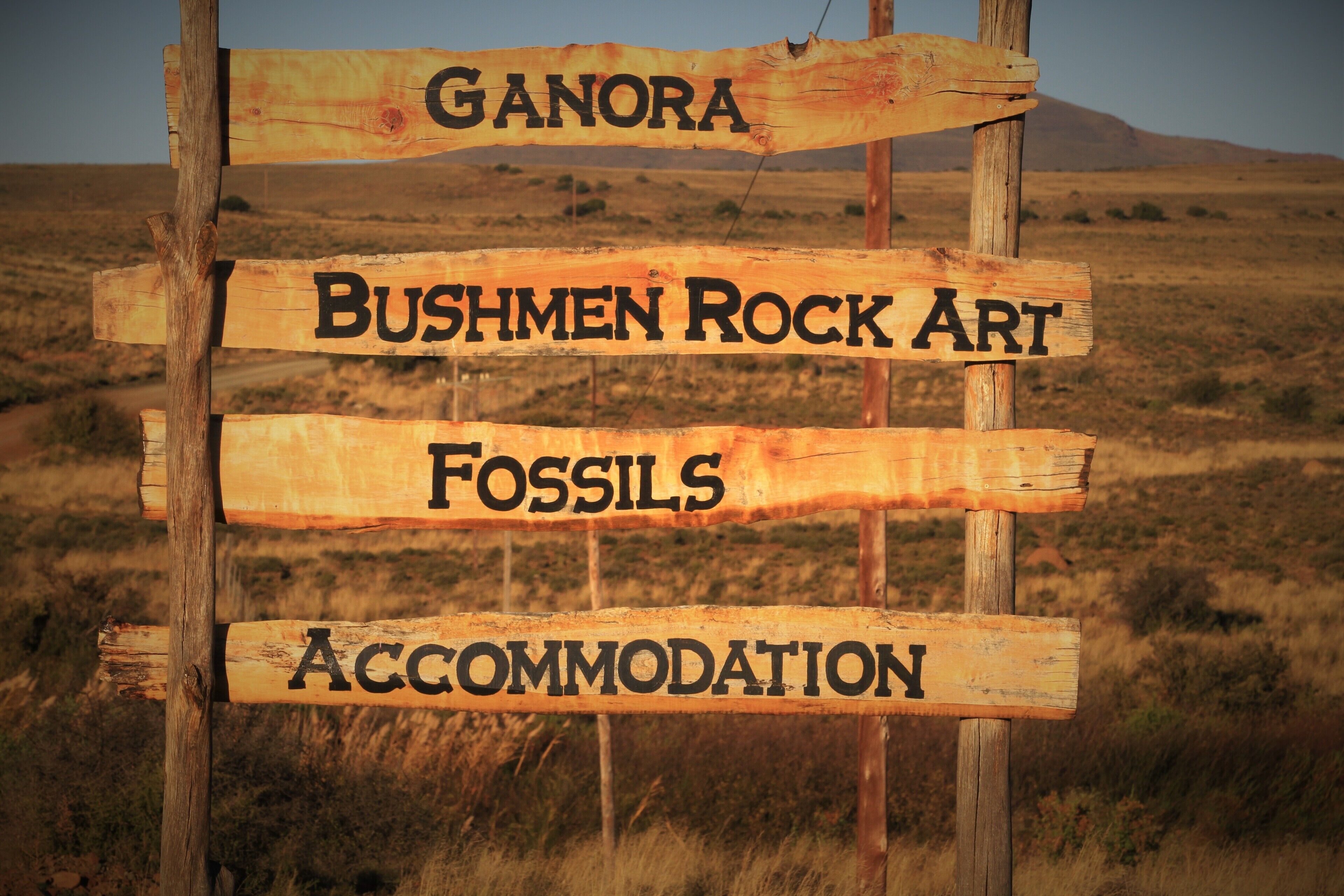 A wooden sign with the words "Ganora," "Bushmen Rock Art," "Fossils," and "Accommodation" indicating points of interest.