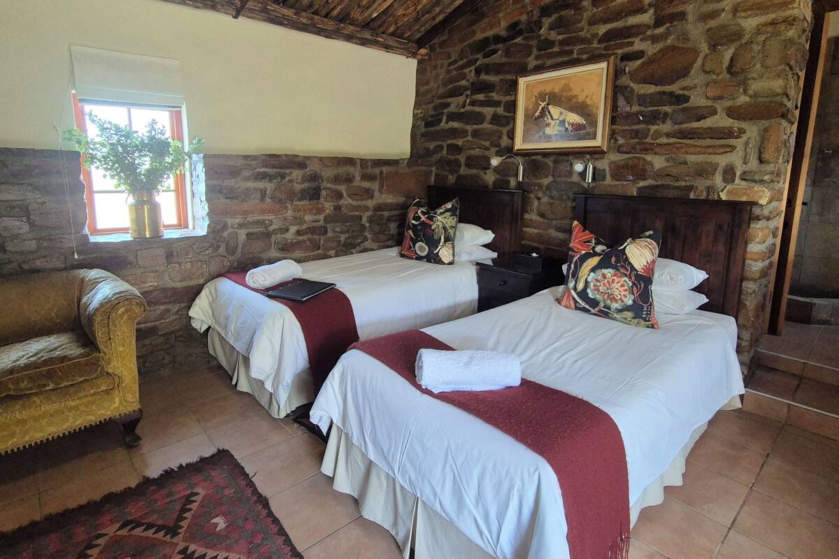 Une chambre rustique avec deux lits, un plafond en bois, des murs en pierre, une fenêtre avec une plante et un tableau encadré accroché au mur.