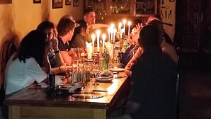 Ein schummrig beleuchteter Raum, in dem eine Gruppe von Personen an einem Tisch mit Kerzen sitzt, umgeben von Werkzeugen und Geräten an den Wänden.