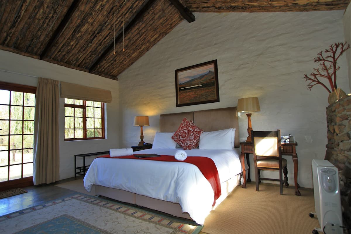 Une chambre avec un grand lit, deux tables de chevet avec des lampes, une chaise, un bureau, un tableau au mur et une fenêtre avec des rideaux.