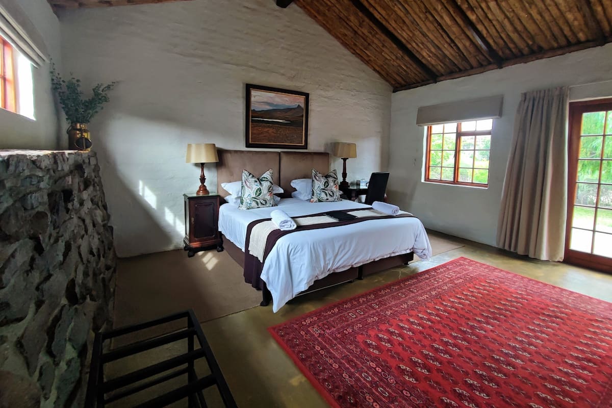 Une chambre à coucher avec un lit, deux tables de chevet avec des lampes, une tête de lit en bois, un tableau au mur, une fenêtre avec des rideaux et un tapis rouge à motifs.
