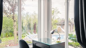 A modern office space with a glass desk and chair, a laptop, and a desk lamp. It features a large window with a view of a backyard with a pool and lounge chairs.