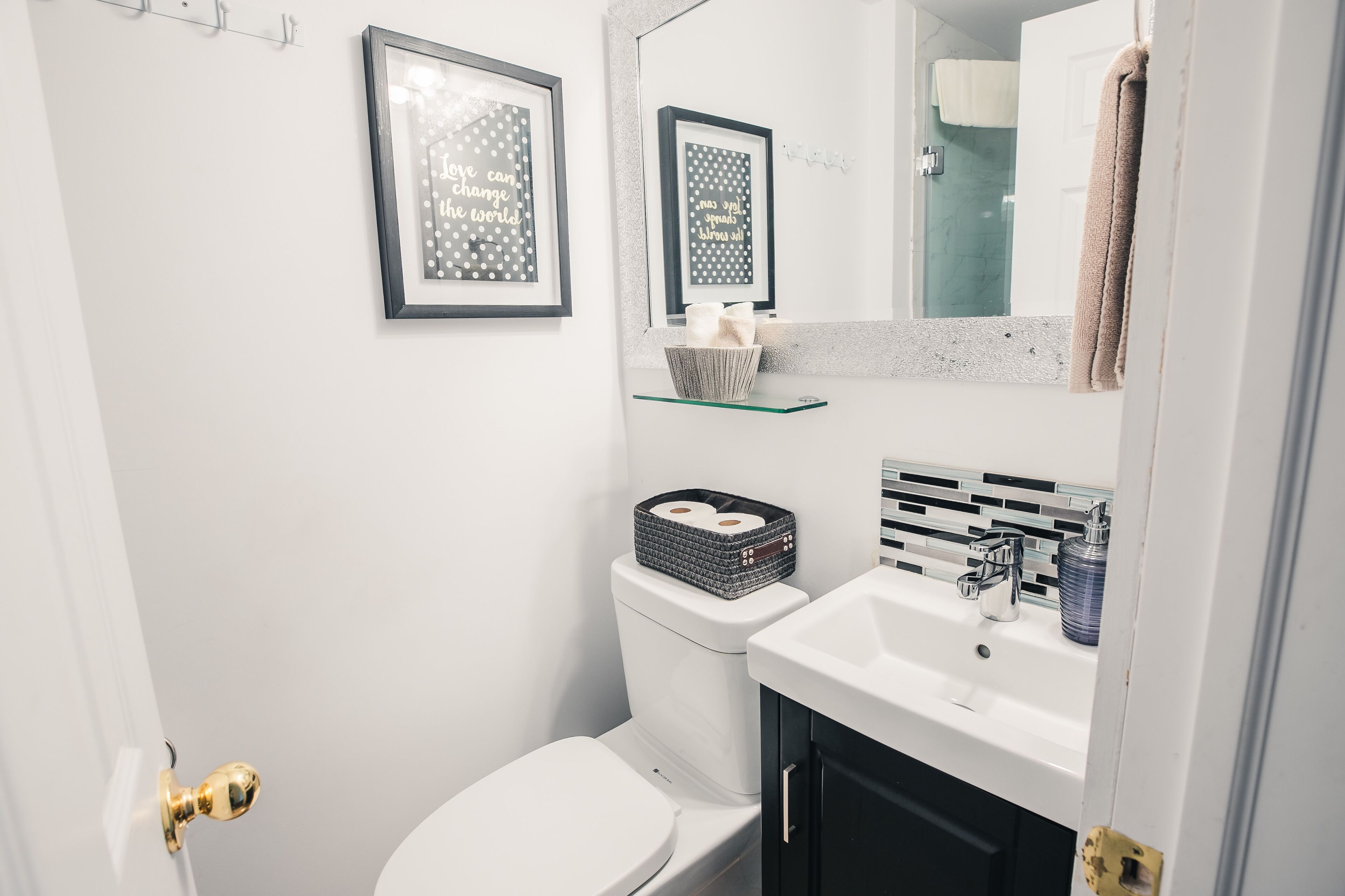 Une salle de bain moderne avec des toilettes blanches, une étagère en verre où sont rangés les articles de toilette et un grand miroir au-dessus du lavabo.